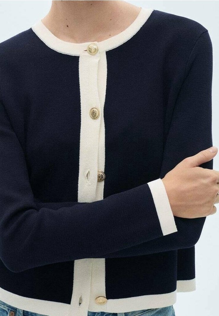 Femme portant un cardigan bleu marine avec une bordure blanche et des boutons dorés, les bras croisés, vue du cou jusqu'aux hanches.
