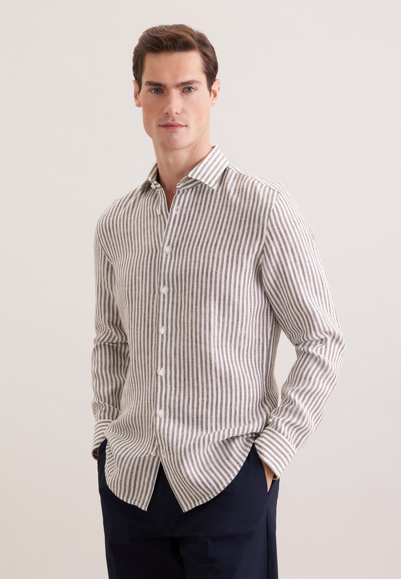 Homme portant une chemise à rayures verticales beige et blanche, un pantalon foncé, les mains dans les poches, debout devant un fond clair uni.