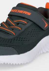 Gros plan sur une chaussure de sport foncée avec des touches orange, une texture en mesh, des lacets rayés, une sangle velcro et une semelle blanche rainurée.