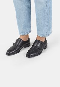 Zapatos de cuero negros con punta redondeada y dos hebillas decorativas. Acabado suave con un ligero brillo. Combinados con jeans de puños enrollados en azul claro.
