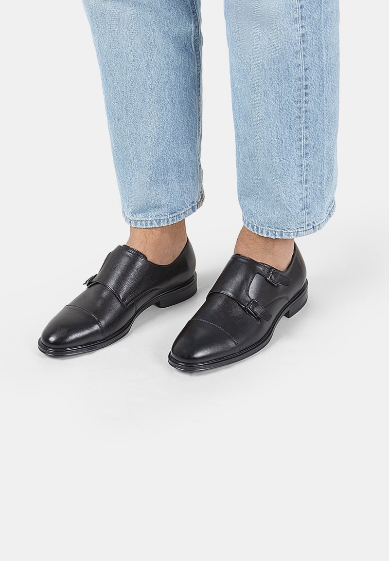 Zapatos de cuero negros con punta redondeada y dos hebillas decorativas. Acabado suave con un ligero brillo. Combinados con jeans de puños enrollados en azul claro.