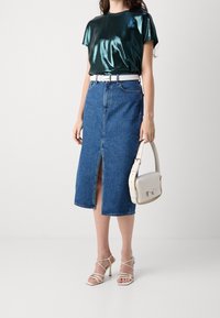 Metallic mörkgrön topp, blå denim midi kjol med framklippning, vit bälte och beige crossbody-väska. Modell bär sandaler med hälar och remmar.