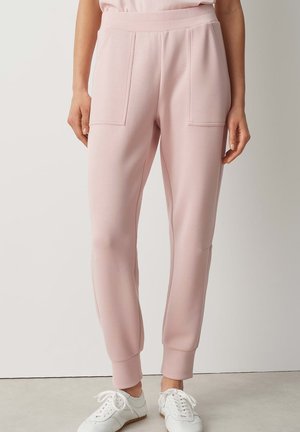 Pantalon de jogging rose clair avec taille élastique et poches plaquées à l'avant, porté avec des baskets blanches à lacets sur un sol gris.