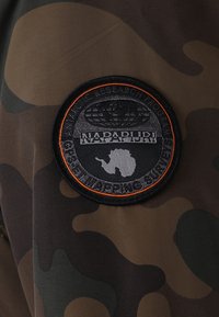 Rund sort patch med orange kant på camouflage stof, der viser en globe, et kort over Antarktis og teksten "NAPAPIJRI Antarctic Research GPS Mapping Surveys."