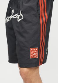 Μαύρο σορτς με κόκκινες κάθετες ρίγες στο πλάι, άσπρα κεντημένα γράμματα κοντά στην τσέπη και κόκκινο έμβλημα με την επιγραφή "Three Stripes One World".