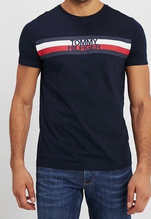 Mężczyzna ubrany w granatowy t-shirt Tommy Hilfiger z białymi, czerwonymi i ciemnoniebieskimi poziomymi pasami na wysokości klatki piersiowej.
