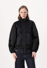 Black, padded jacket with a high collar and elastic hem. Features a front closure with buttons and wide, gathered sleeves.