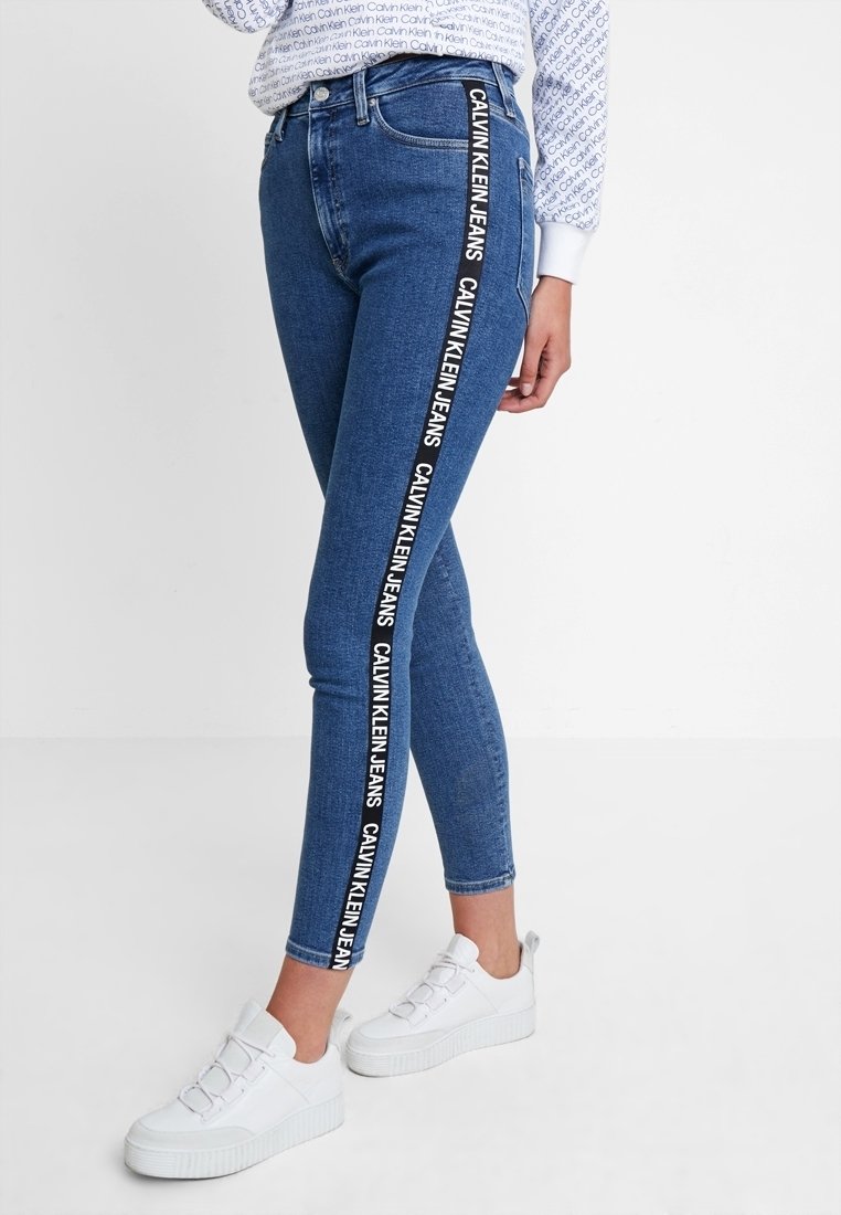 Jeans ajustados de mezclilla en azul, con rayas laterales negras que llevan el texto "CALVIN KLEIN JEANS". Combinados con zapatillas de plataforma blancas.