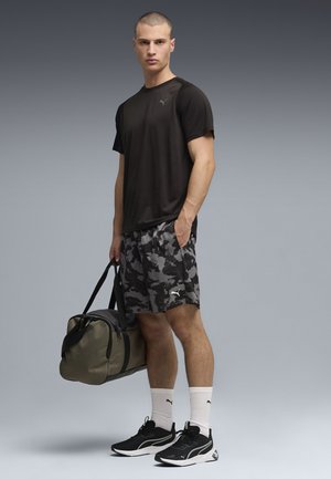 Mężczyzna ubrany w czarną sportową koszulkę Puma, kamuflażowe szorty, białe skarpetki Puma, czarne sneakersy, trzymający oliwkową torbę sportową, stojący na szarym tle.