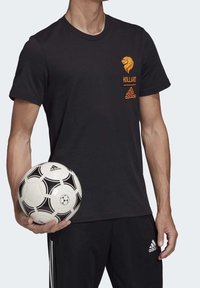 Černé tričko s krátkým rukávem s grafickým motivem oranžového lva a textem "Holland"; v kombinaci s černými sportovními kalhotami, které drží černobílý fotbalový míč.