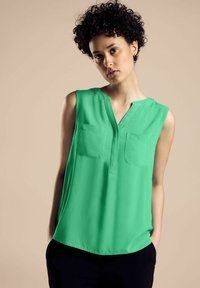 Väri vihreä, ilman hihoja oleva paita kevyestä kankaasta V-aukkolla, kahdella rintataskulla ja pyöristetyllä helmallä, yhdistetty tummiin housuihin.