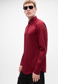 Giovane uomo con occhiali da sole neri a forma rettangolare e una camicia a maniche lunghe borgogna aderente, in piedi su uno sfondo bianco uniforme.