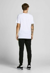 Hvid t-shirt med korte ærmer, parret med sorte joggers med et grønt og blåt mønster på siden, samt sorte træningssko.