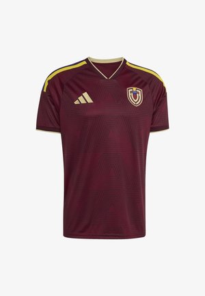 Camiseta de fútbol color marrón con un patrón geométrico, detalles dorados y escote en V. Presenta rayas azul y amarilla en los hombros y un emblema en el pecho.