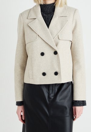 Femme portant une veste en laine beige à double boutonnage sur un pull gris foncé et une jupe en cuir noire, montrée des épaules à la mi-cuisse.