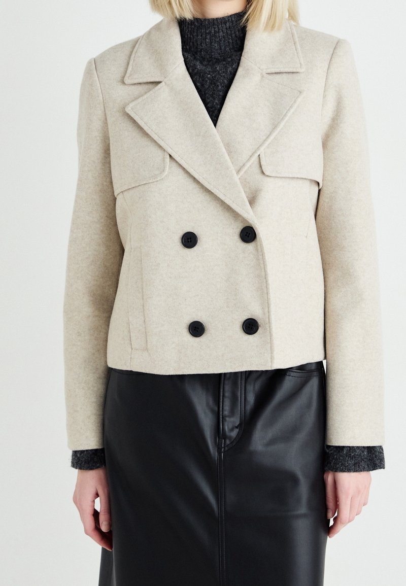 Femme portant une veste en laine beige à double boutonnage sur un pull gris foncé et une jupe en cuir noire, montrée des épaules à la mi-cuisse.