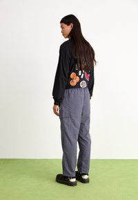 Schwarzes Langarmshirt mit bunten Obstgrafiken auf dem Rücken, kombiniert mit grauen Cargo-Hosen und schwarzen Schuhen mit weißen Akzenten.