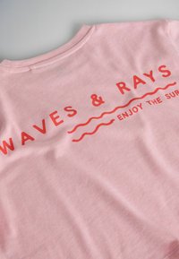 Tissu rose avec un texte rouge "WAVES & RAYS" au-dessus de lignes ondulées et un texte plus petit "ENJOY THE SURF" sur un t-shirt.