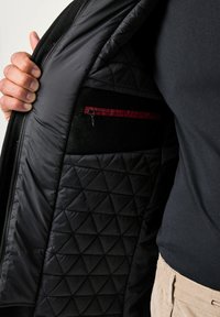 Mano che tiene aperto un giubbotto nero trapuntato mostrando una tasca interna con zip bordata di rosso scuro, indossato sopra una maglietta nera e pantaloni beige.