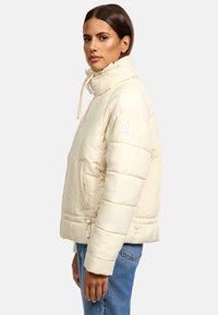 Cream puffer jacket featuring a high collar, zippered front pockets, and adjustable drawstrings. Smooth, quilted texture with a boxy fit.