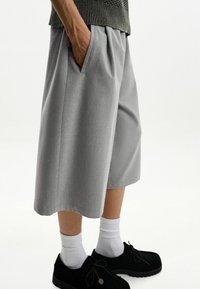 Pantaloni corti grigi su misura con una vestibilità rilassata, dotati di tasche laterali e una superficie liscia. Indossati con calzini bianchi e scarpe nere.