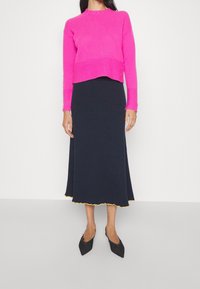 Fuksjowy krótki sweter o prążkowanej strukturze, zestawiony z granatową, prążkowaną midi spódnicą z falowanym dołem i żółtym wykończeniem. Czarne spiczaste buty.