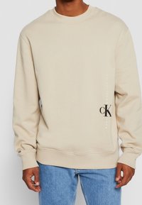 Sweatshirt beige à col rond avec des poignets et un ourlet côtelés. Présente un design de logo noir sur le côté, fabriqué à partir d'un mélange de coton doux.