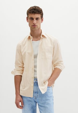 Ung mand iført en beige stribet skjorte med knapper over en hvid tanktop og lyseblå jeans, stående med den ene hånd i lommen.