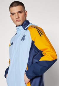 Svijetloplava, mornarsko plava i narančasta jakna od vjetra s patentnim zatvaračem, ima rebraste manžete i logo na prsima, čime dodaje kontrast i sportski dizajn.