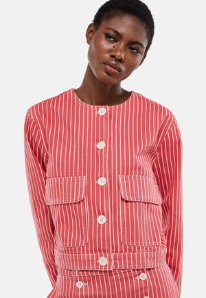 Femme portant une veste boutonnée à rayures verticales rouges et blanches avec deux poches à rabat frontales, regardant légèrement vers sa gauche.