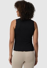 Czarny bezrękawnik z falbanami koronkowymi na ramionach, dopasowany, prążkowany teksturą i wykończeniem. Nosi się go z beżowymi dopasowanymi legginsami.