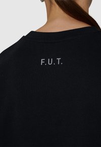 Svart bomullströja med rund halsringning och bokstäverna "F.U.T." tryckta i grått på övre delen av ryggen. Mjuk textur, avslappnad design.