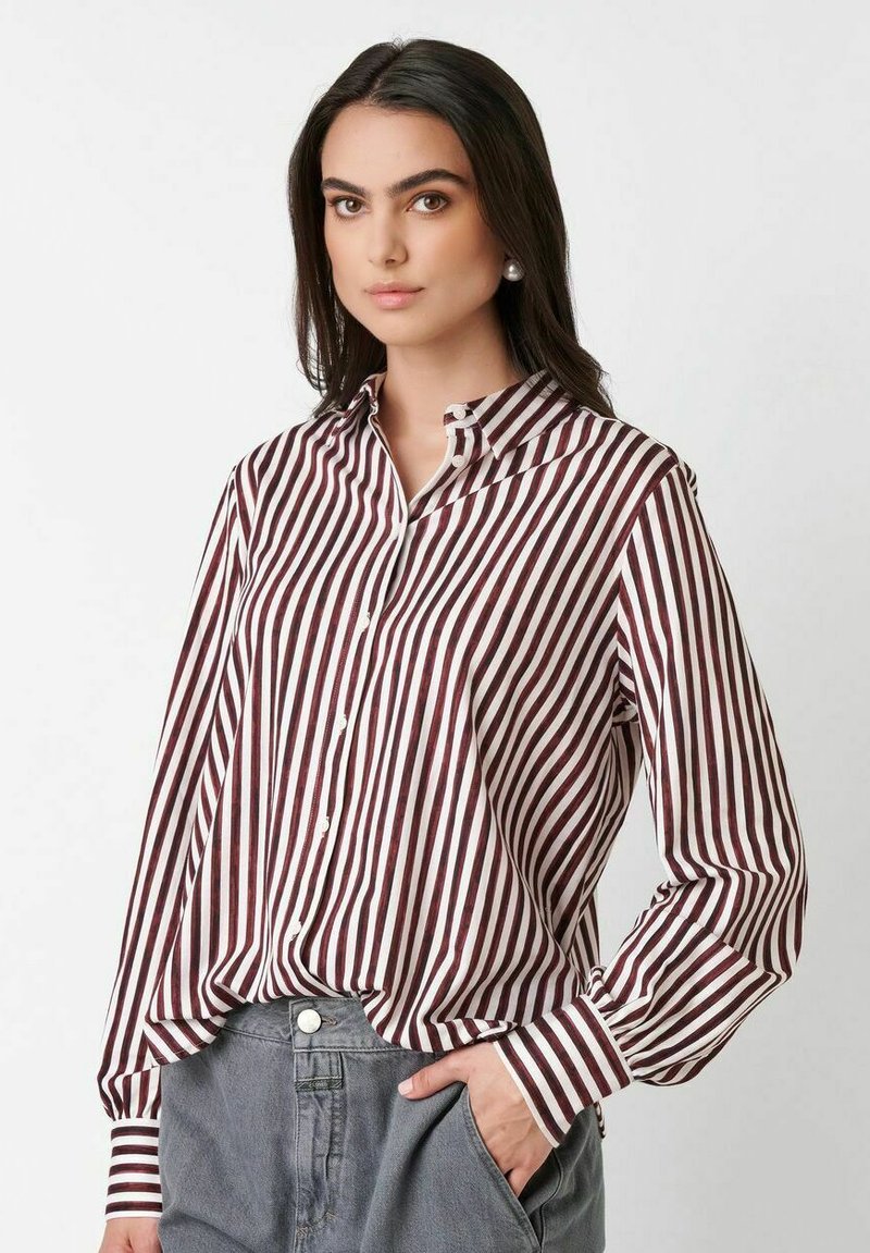 Young woman with long dark hair wearing pearl earrings, a maroon and white vertical striped shirt, and gray pants with one hand in pocket.