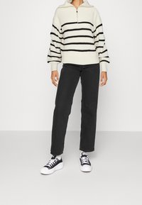 Pull en maille blanche avec des rayures horizontales noires, col zippé, manches oversize ; associé à un jean droit noir et des baskets noires.