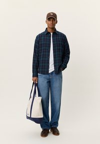 Καρό πουκάμισο από φλανέλ σε ναυτικό μπλε και πράσινο, λευκό T-shirt, φαρδιά τζιν παντελόνια, καφέ καπέλο, κρατώντας διχρωμη τσάντα ώμου με ναυτικές λεπτομέρειες.