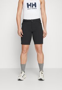 Pantalones cortos negros hechos de una tela elástica, con un frente plano, cierre de botón y bolsillos. Combinados con calcetines de punto grises y zapatillas deportivas.