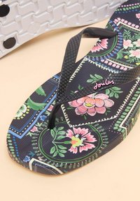 Flip-flop with a floral pattern in pink and green on a black background. Black textured straps, featuring a logo in pink. White textured sole.
