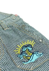 Tissu en denim à rayures bleues avec une poche arrière ornée d'un soleil, d'un palmier et de motifs de vagues brodés en jaune vif et bleu. Accents métalliques visibles.