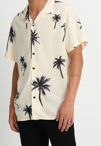 Camisa de manga corta en tejido crema con un estampado de palmeras en negro, con cuello y botones negros en la parte delantera.