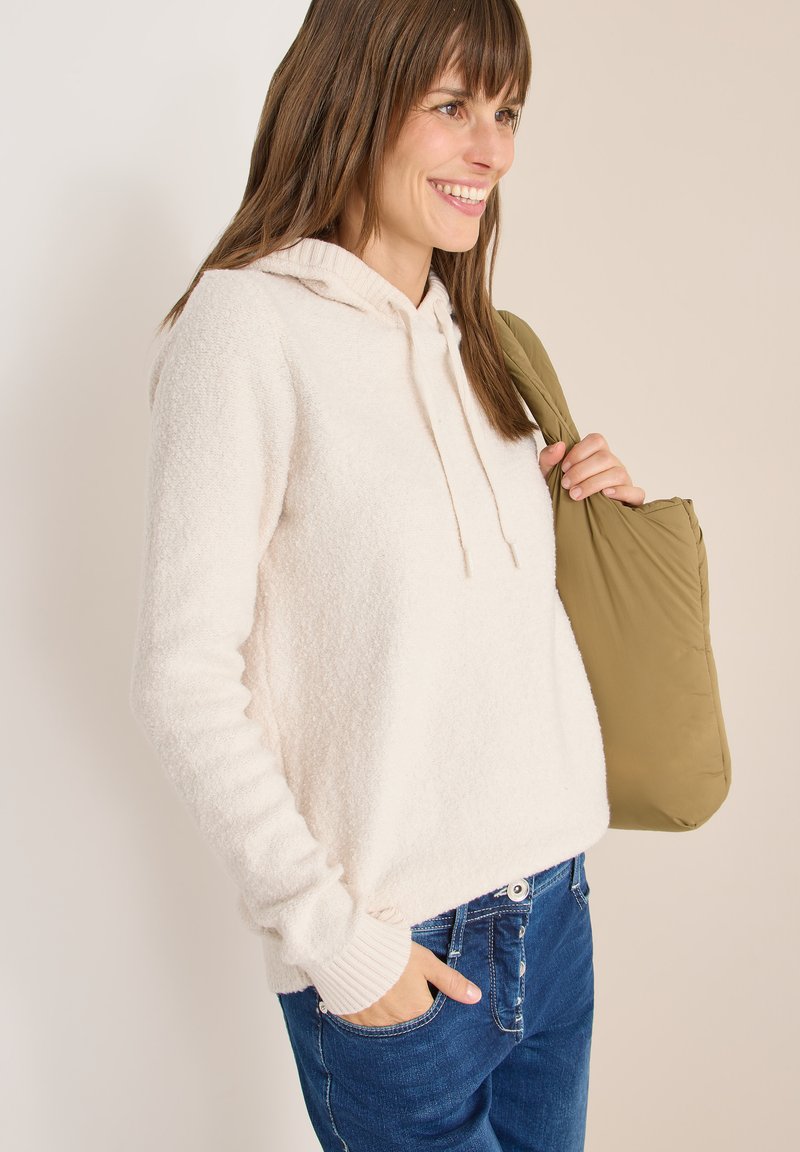 Hoodie en polaire beige clair avec un cordon de serrage, des poignets côtelés et une coupe décontractée, associé à un jean bleu foncé et un sac vert olive.