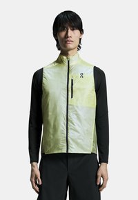 Letvægts gul vest med høj krave, lynlåslukning foran og sorte detaljer. Båret over en sort langærmet skjorte. Slank, strømlinet design.