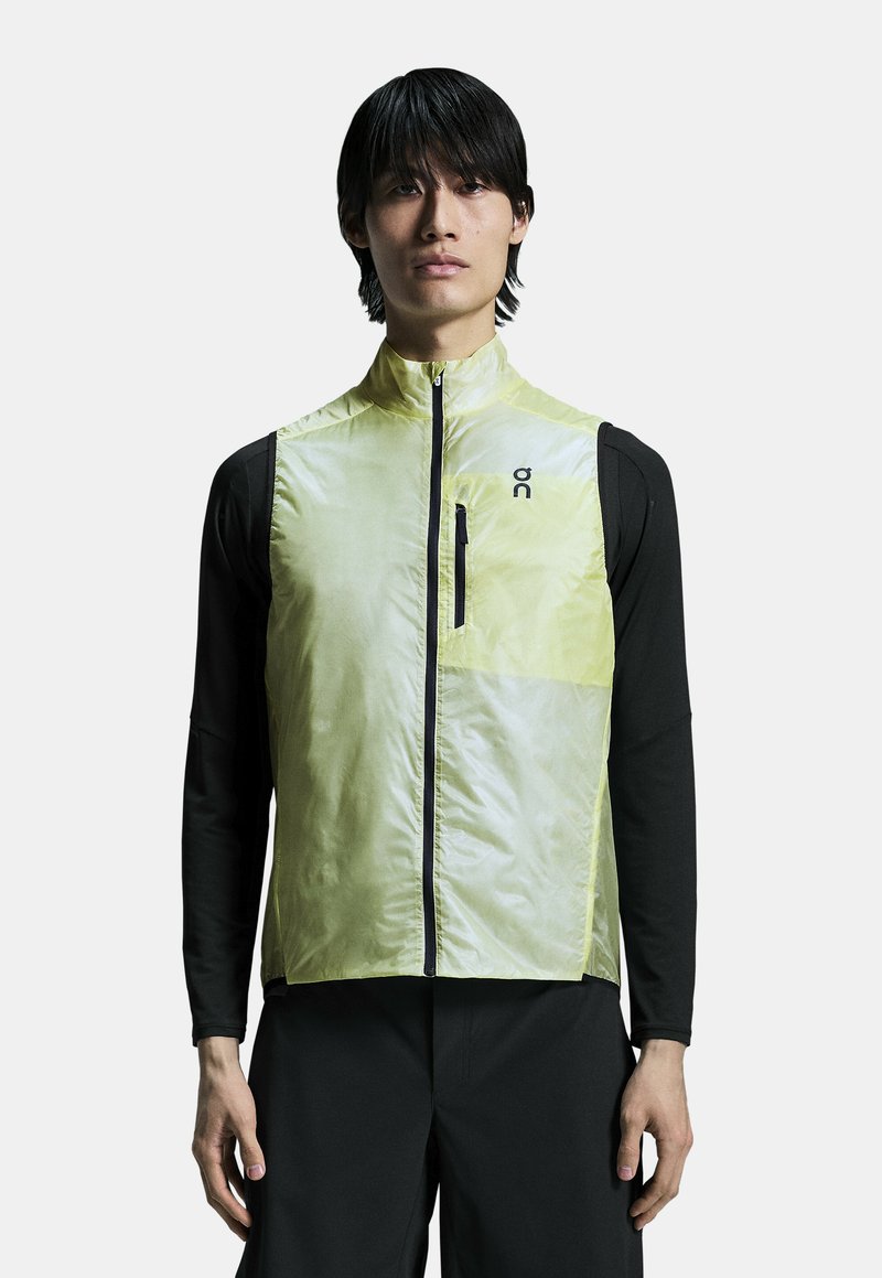 Lightweight yellow vest with a high collar, zippered front, and black accents. Worn over a black long-sleeve shirt. Sleek, streamlined design.