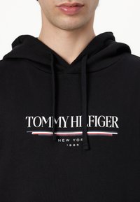 Svart huvtröja i bomull med en stor vit "TOMMY HILFIGER"-logotyp och röda och blå randar under.