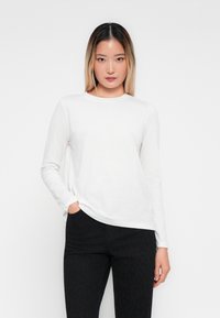 Young woman with straight black-to-blonde hair wearing a white long-sleeve shirt and black pants, standing with one hand in pocket.