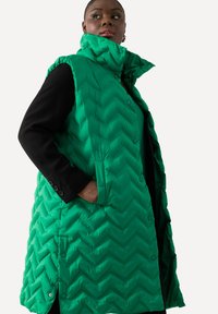 Grön vadderad väst med ett chevronmönster, hög krage och sidofickor. Den har ett knappstängning och en mjuk, glänsande yta.