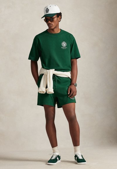 Grønn bomullsjakke og shorts med logo, kremfarget genser bundet rundt livet, grønn caps, solbriller og grønne joggesko på en nøytral bakgrunn.