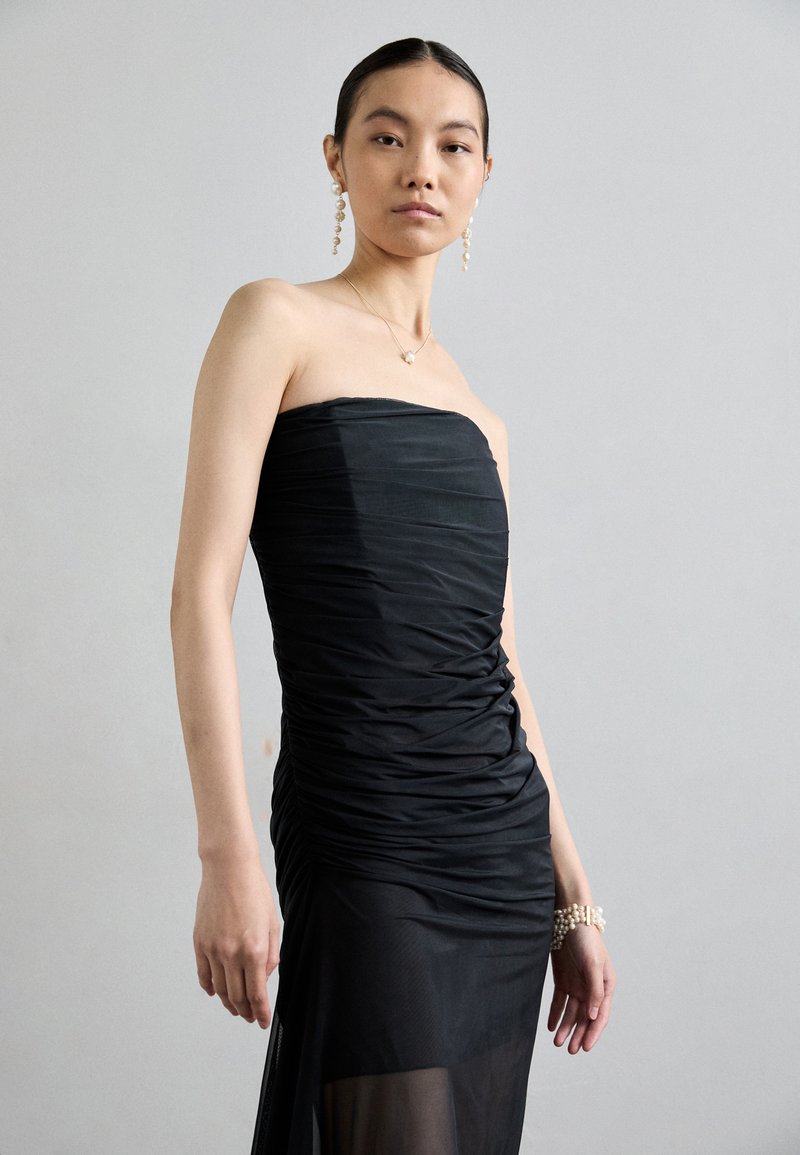 Abito nero senza spalline con texture arricciata, silhouette fluida, sovrapposizione di tessuto trasparente, indossato con orecchini e bracciale di perle.