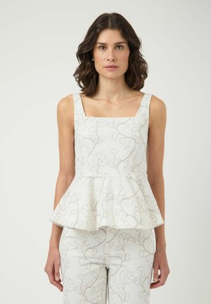Femme aux cheveux foncés mi-longs portant un top peplum blanc sans manches et un pantalon assorti à motif floral clair, debout devant un fond uni.