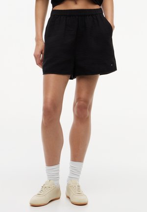 Donna con pantaloncini neri a vita elastica, calzini bianchi a costine e sneakers beige, in piedi con le mani nelle tasche contro uno sfondo bianco.