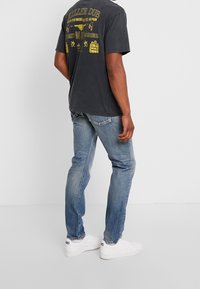 Černé tričko s žlutým grafickým potiskem na zádech, v kombinaci s světlými modrými džíny a bílými teniskami. Jednoduchý design a volný střih.